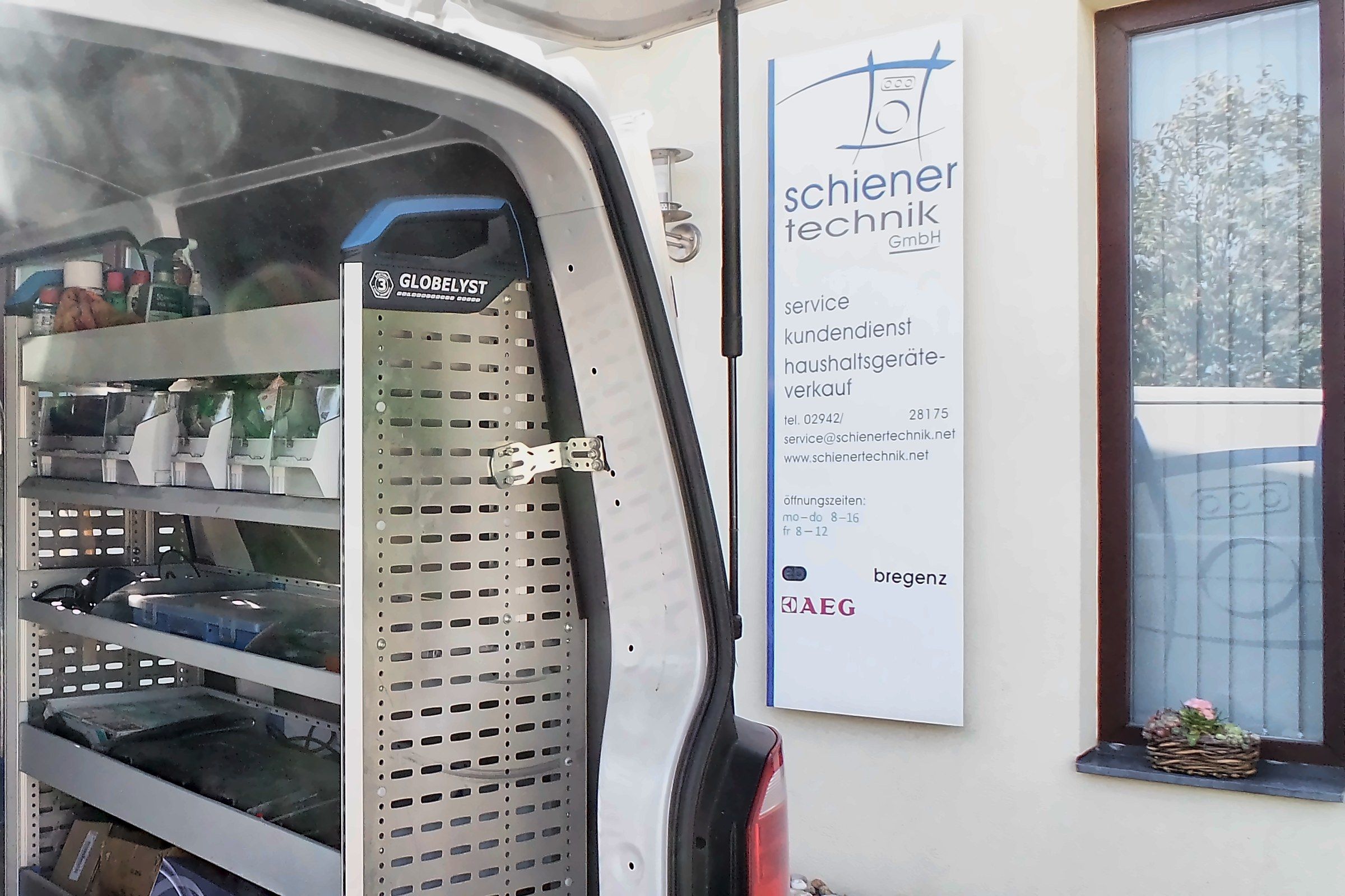 Geöffnete Hecktür eines Servicefahrzeugs mit Regaleinbau, daneben das Firmenschild am Gebäude