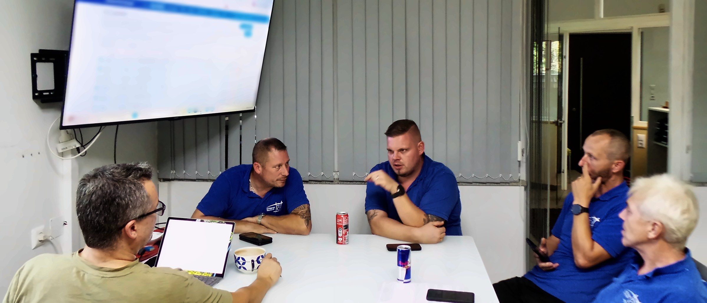 Team von schiener technik im Gespräch am Besprechungstisch, die meisten in blauen Firmen-Poloshirts